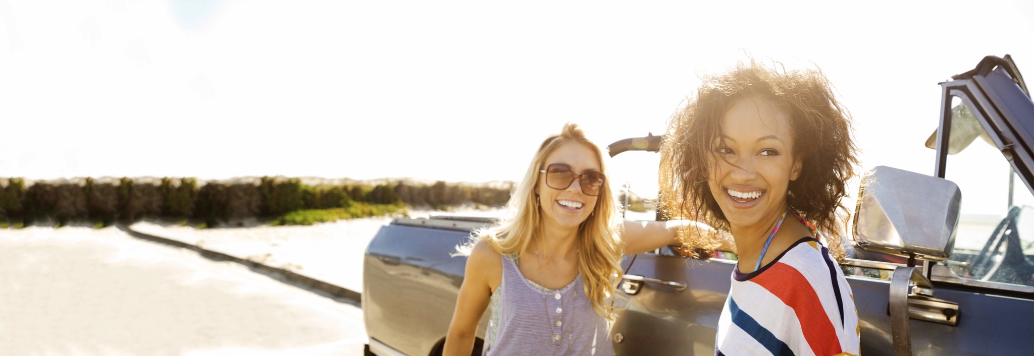 Two women smiling, one with smooth, dry hair and the other one with curly, dry hair, next to a car.