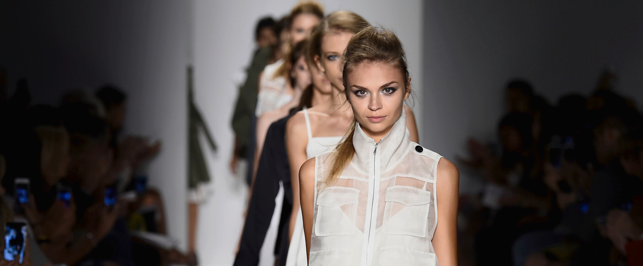 A line of models on the runway with a model wearing a white sleeveless jacket with pockets  