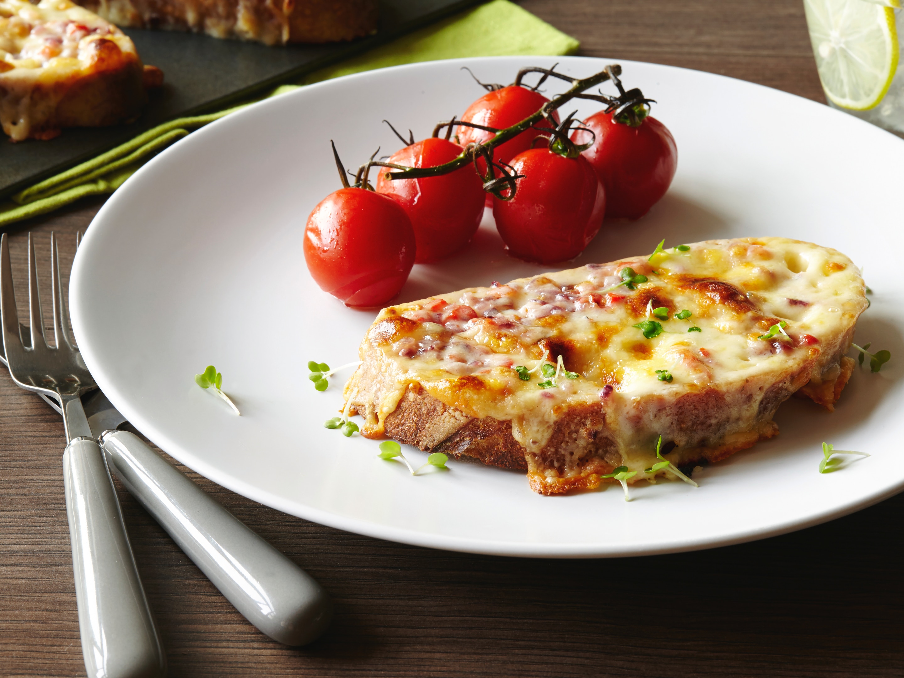 Welsh rarebit beside a vine of cherry tomatoes. on a white plate. White handled cutlery is in the foreground with toasted bread in the background.