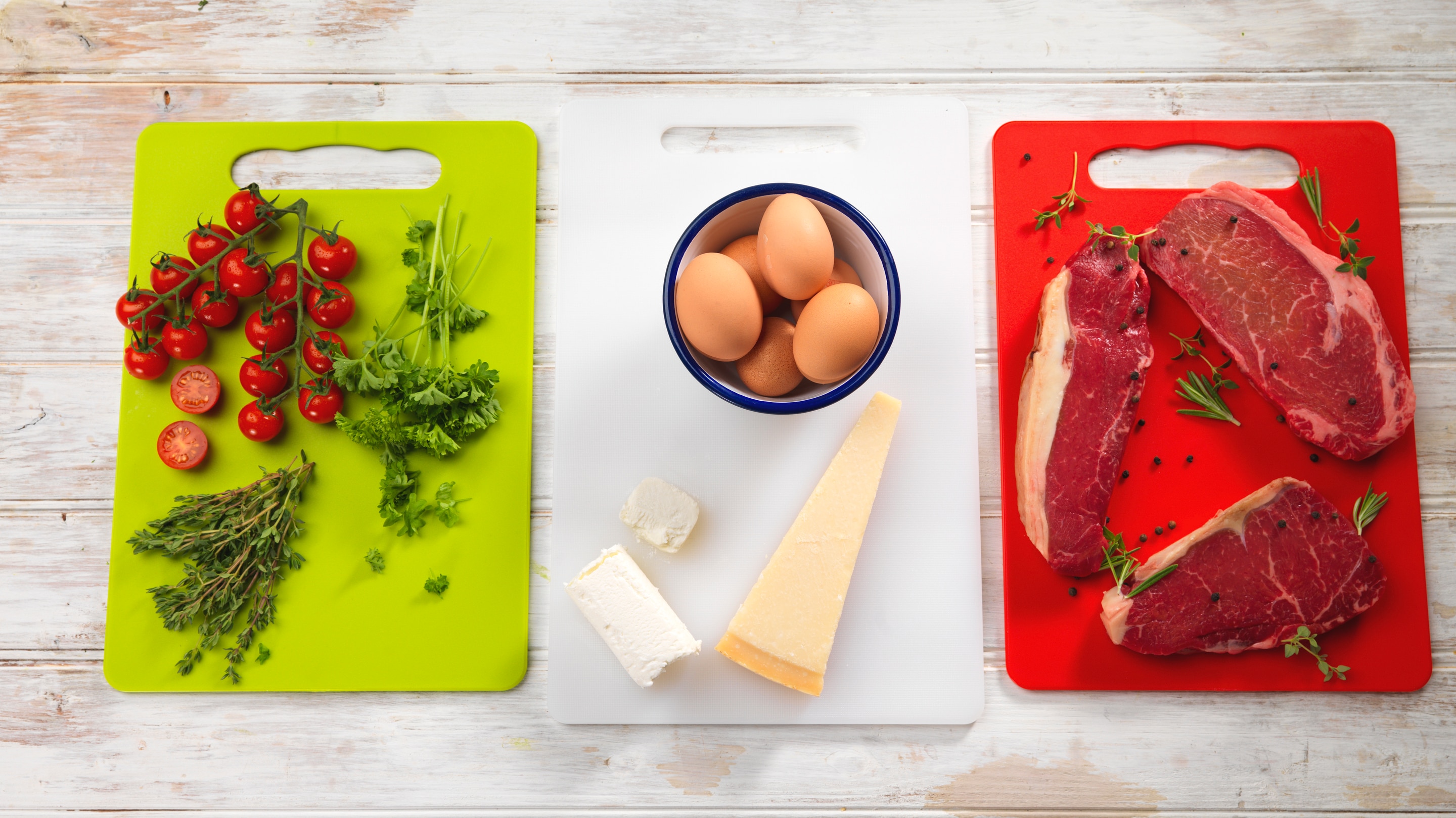 Tomatoes, parsely & rosemary on green chopping board, eggs & cheese on white chopping board and steak on red chopping board