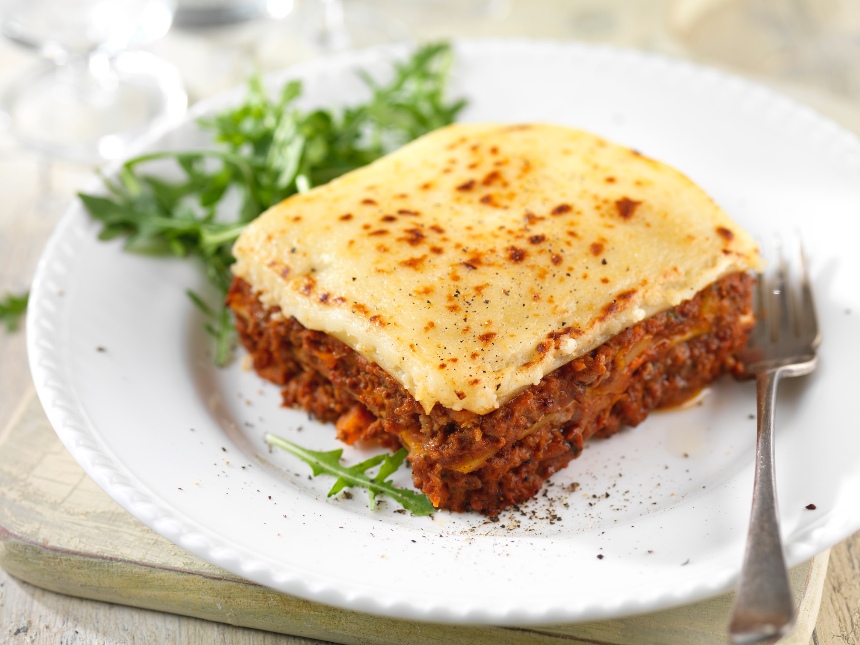 A lasagne served in a white plate and a silver fork placed beside. It is served with a few salad leaves.