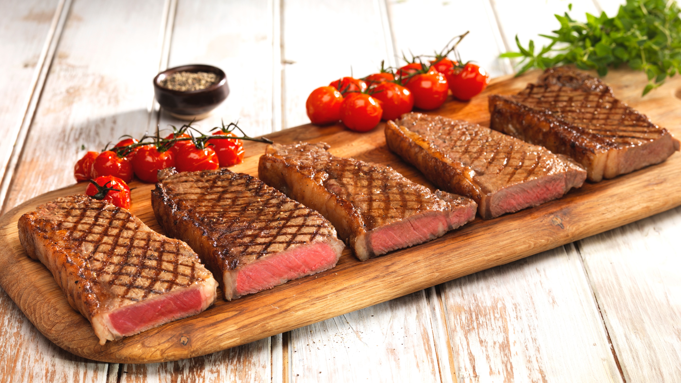 Grilled steak pieces placed on a wooden surface with bunch of cherry tomatoes and green leafy salad.  