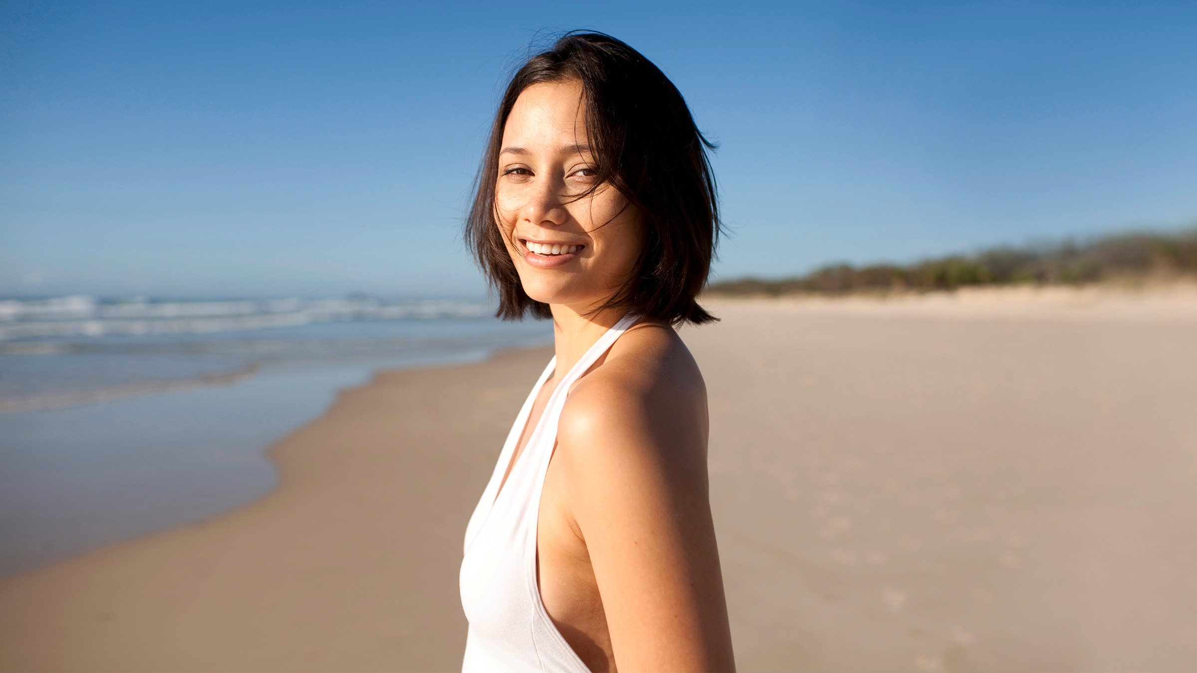 A person is shown standing on a beach and smiling. They are turned to the side and facing the camera.