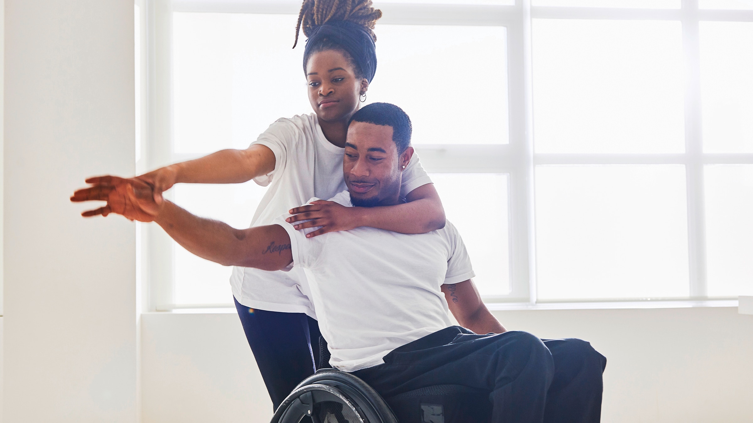 A woman helping a man in a wheelchair stretch his arm indoors.