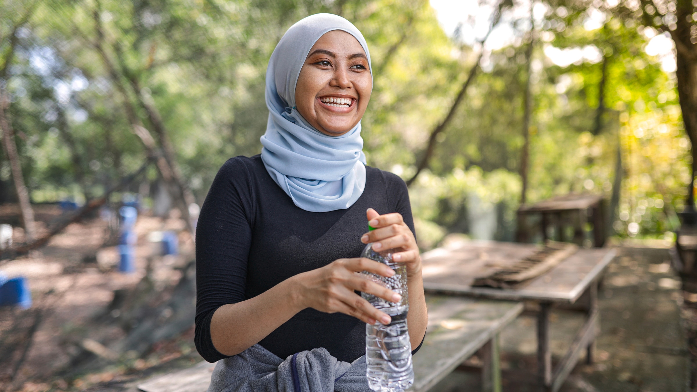 A person is shown smiling and holding a water bottle. They are outside and tables can be seen in the background.