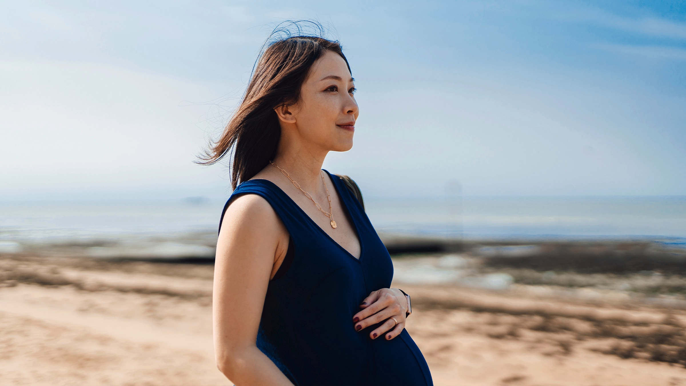 A side shot of a woman at the beach. She has her hand on top of her stomach and appears to be pregnant.