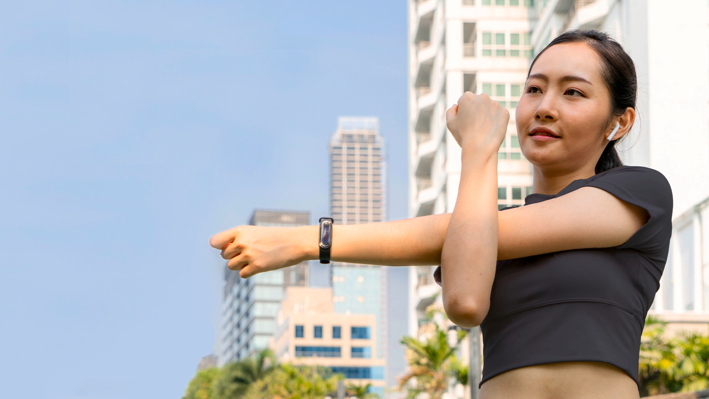 A person is shown outside stretching their arm. They are wearing wireless ear buds and buildings can be seen in the background. 