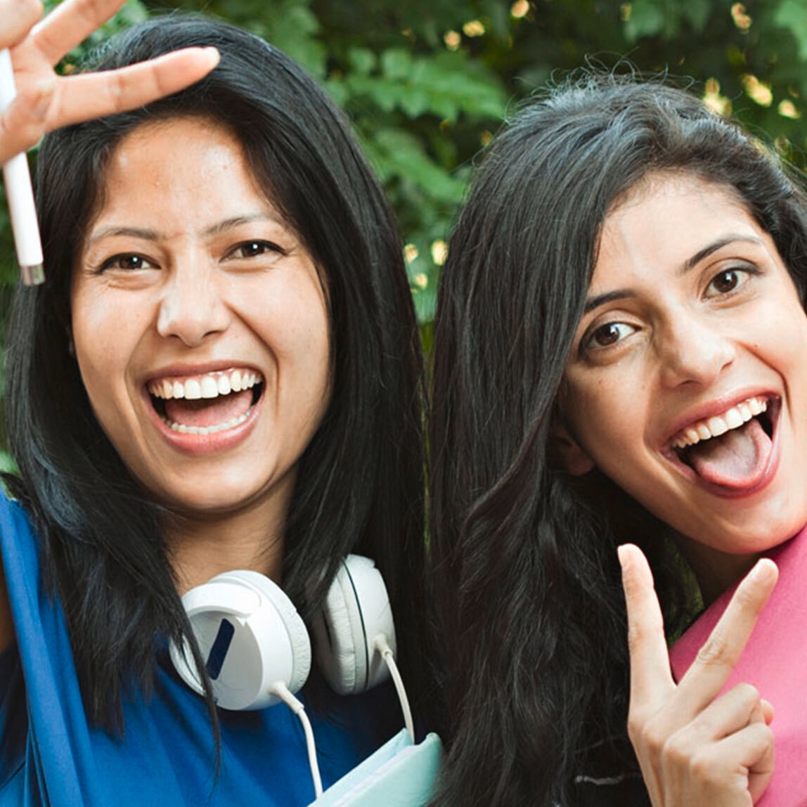 Two women laughing and making two-fingered peace gesture at the camera. One of the girls is wearing large headphones and holding a school notebook.