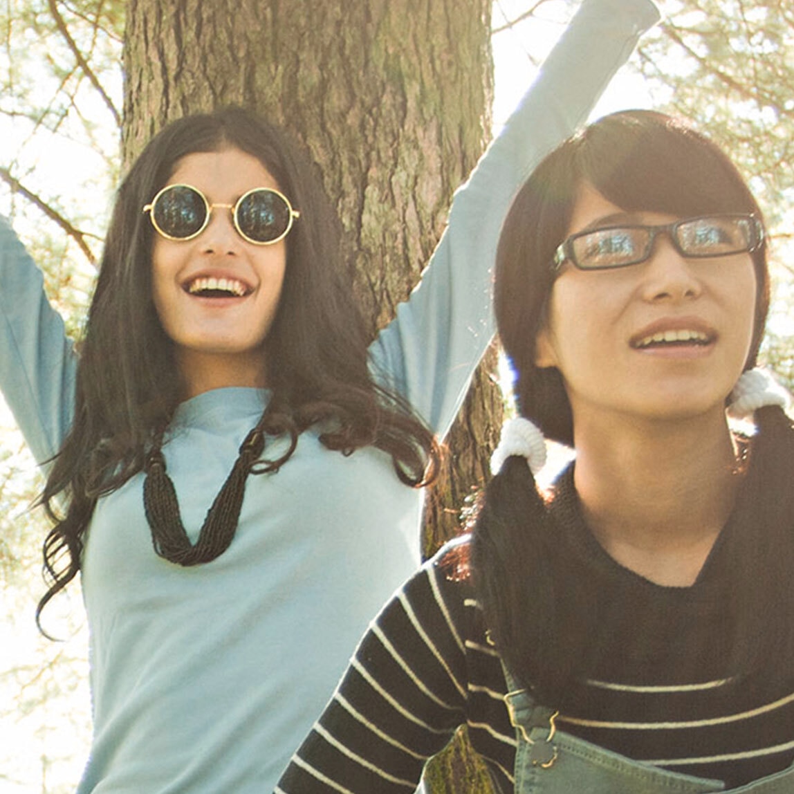 Three young women with long, thick black hair standing around looking cheerful in a sunny woodland.  