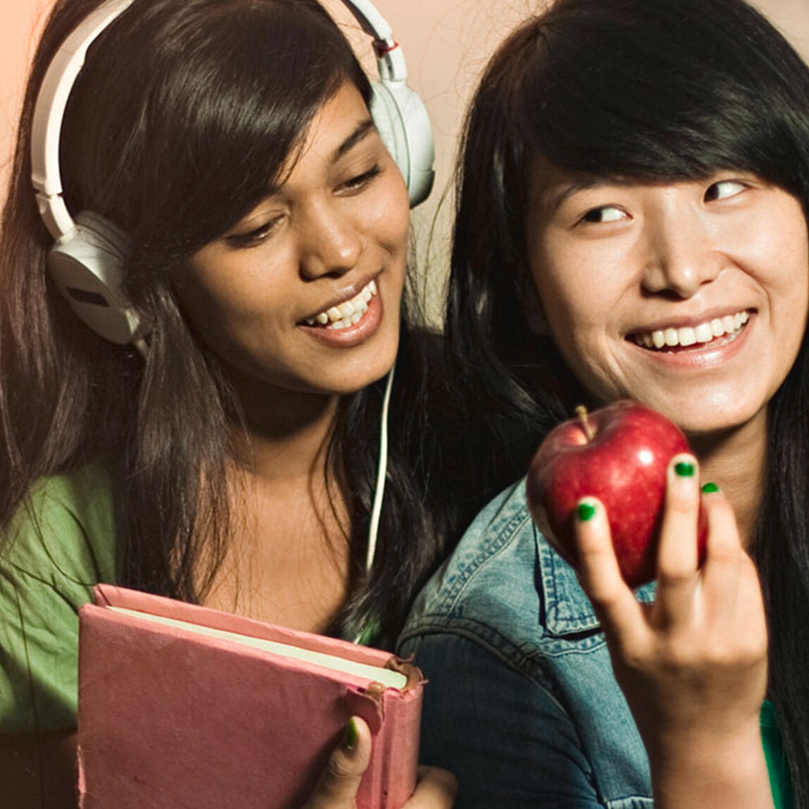 Two women with thick black hair. One is holding a red apple, the other is wearing large headphones and looking over the first's shoulder.