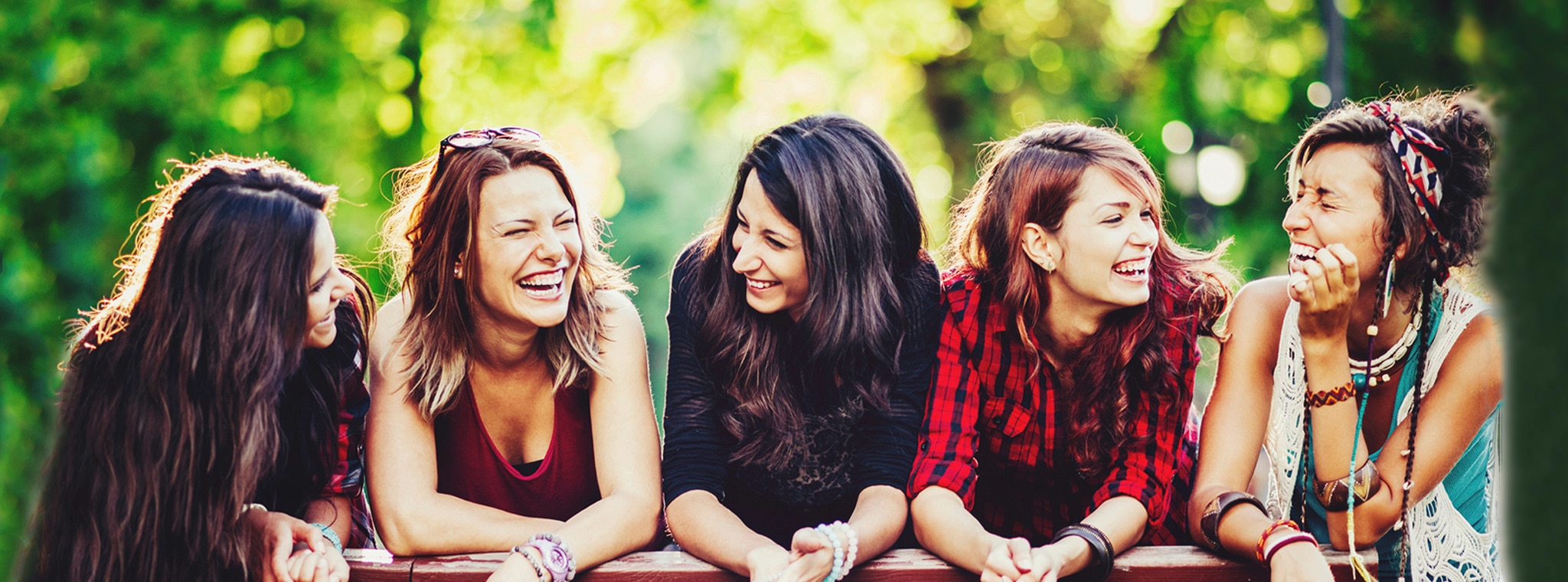 A group of girlfriends with straight and curly hair laughing together on a bridge by a lake.