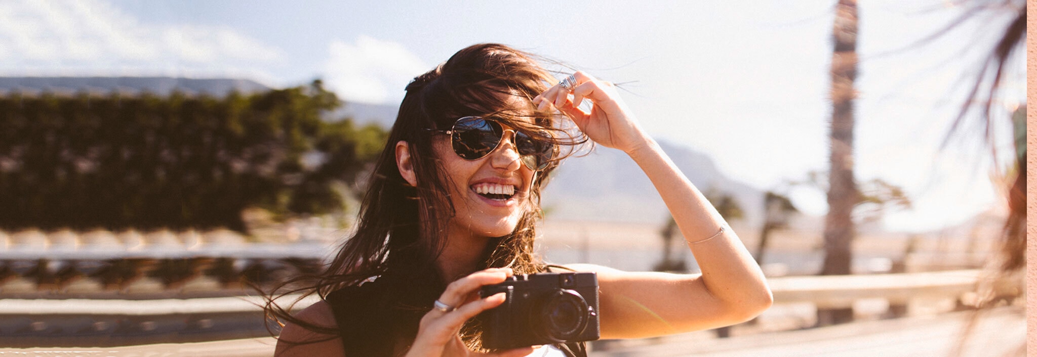 A woman standing on a beach and holding a camera, laughing as the wind blows her brown hair into her face.