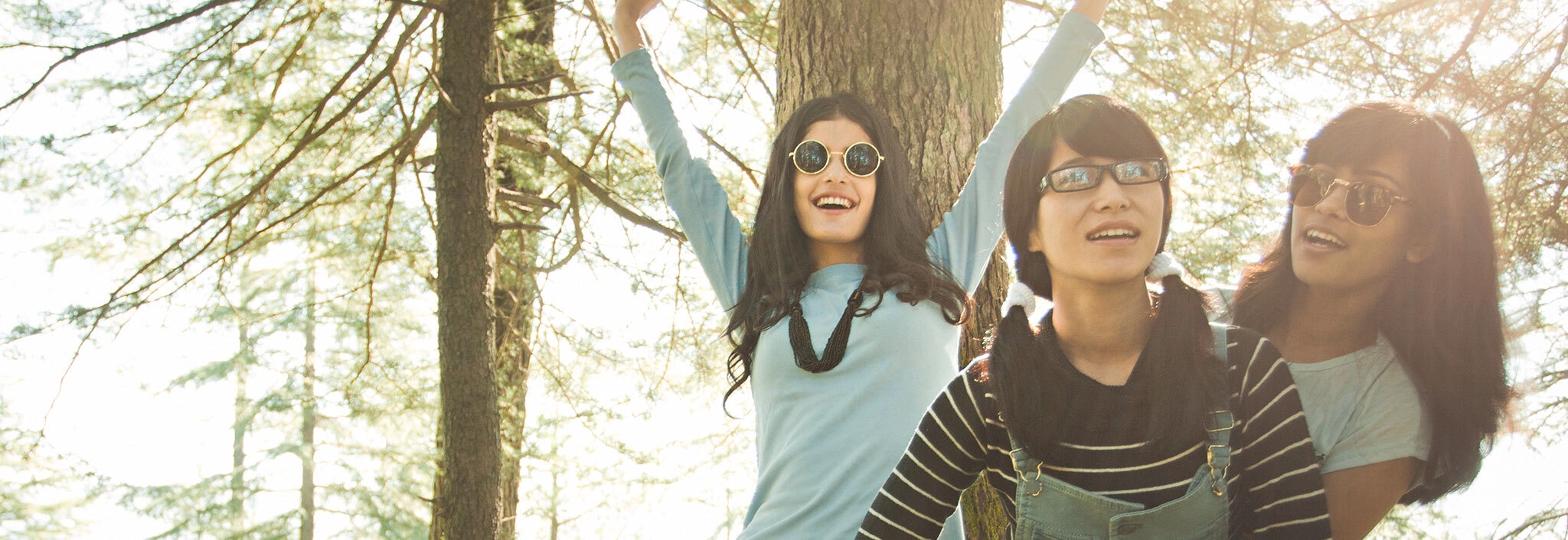Three young women with long, thick black hair standing around looking cheerful in a sunny woodland
