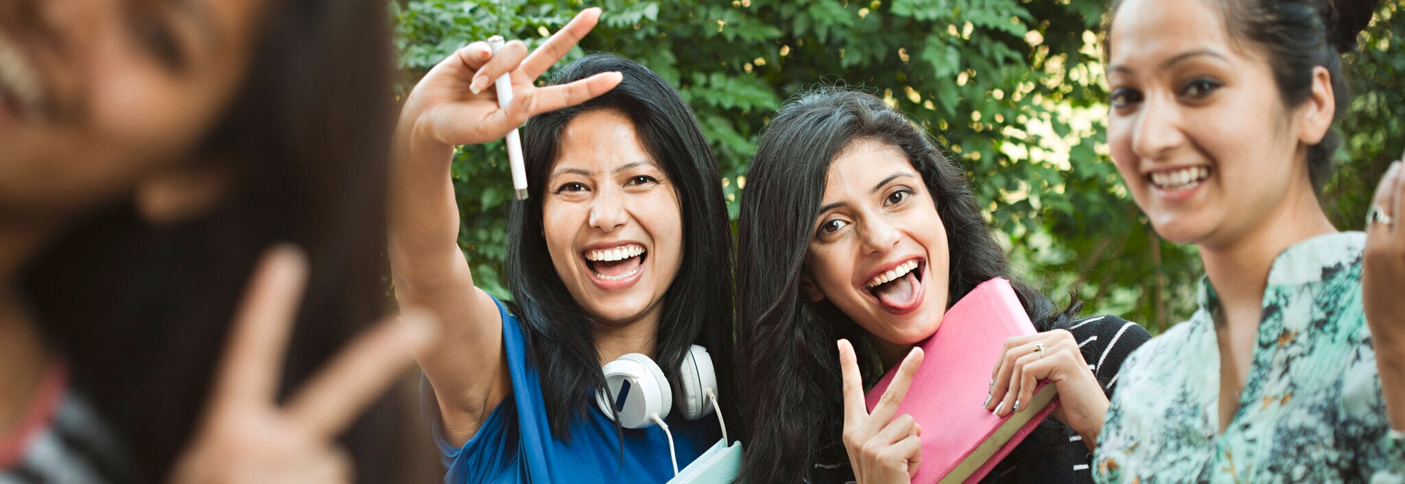 Four women laughing and making two fingered peace gestures towards the camera. One wears large headphones and holds a school notebook
