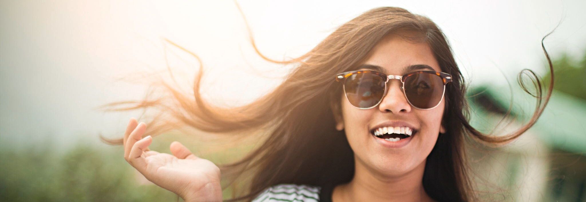 A smiling woman wearing sunglasses, her wavy black hair blowing gently in the wind.