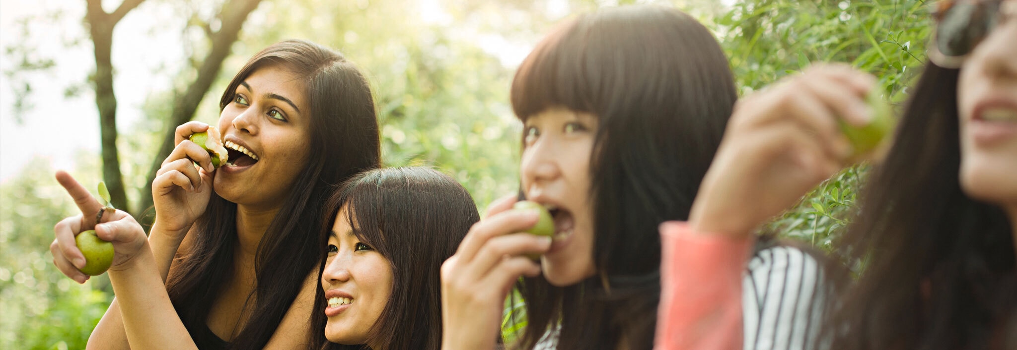 Four women, all with thick black hair, eating apples and pointing towards something in the distance.