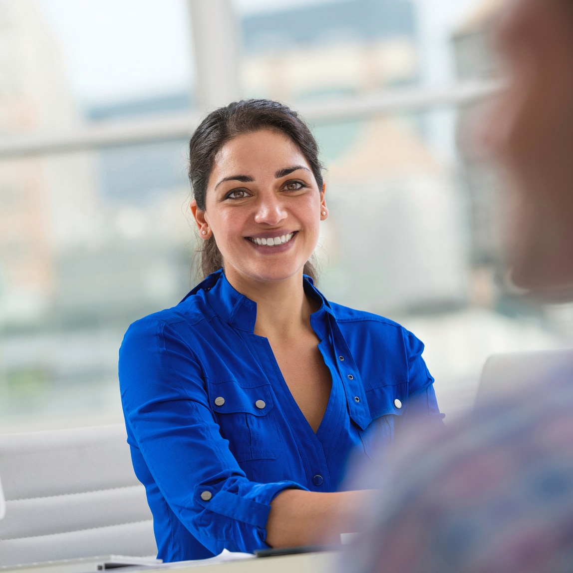 A smiling woman in a bright blue shirt sitting next to a desktop computer monitor.