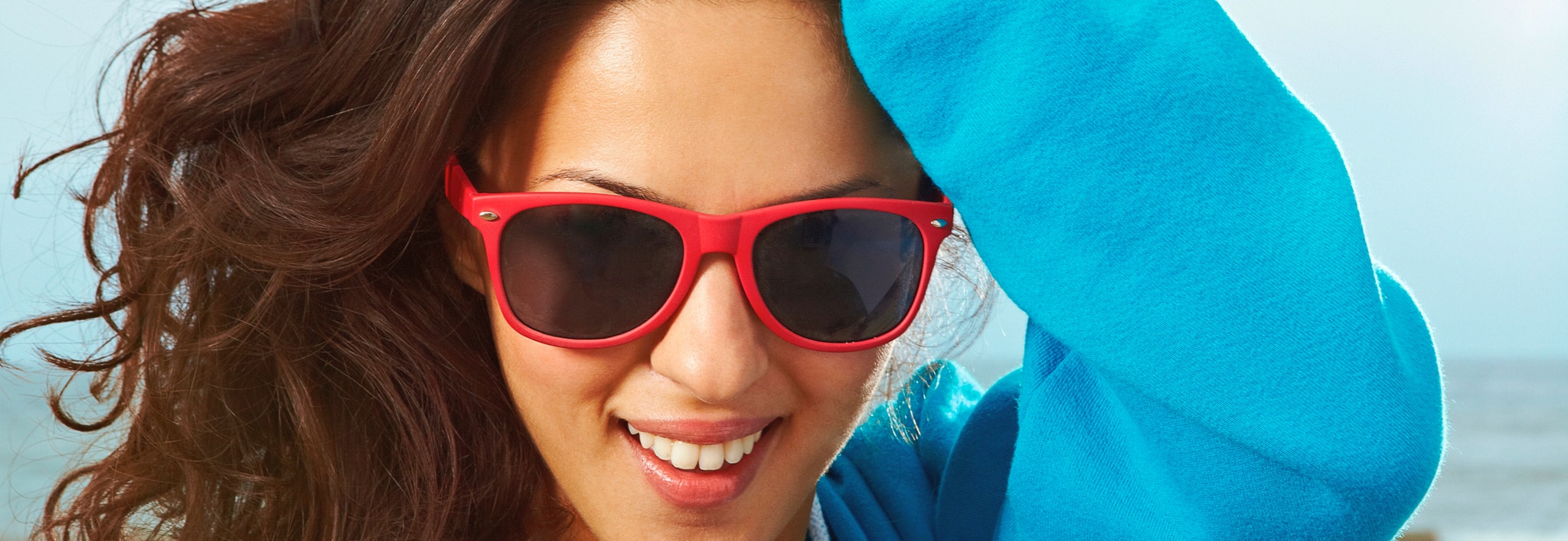 A smiling woman wearing red sunglasses, running a hand through her thick, wavy, brown hair. 