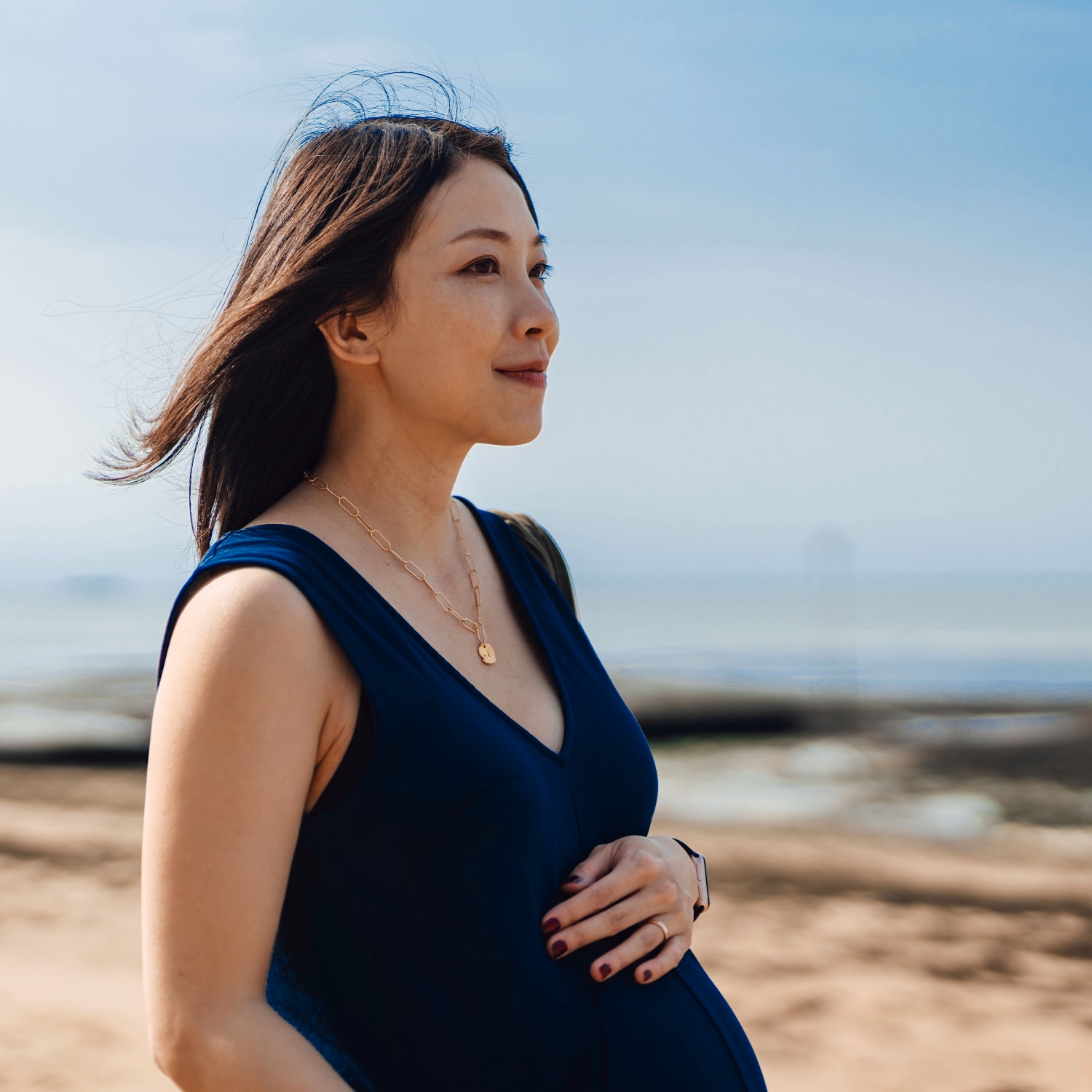 A side shot of a person is shown as the beach is shown in the background. They are holding the top of their stomach as they seem to be pregnant. 