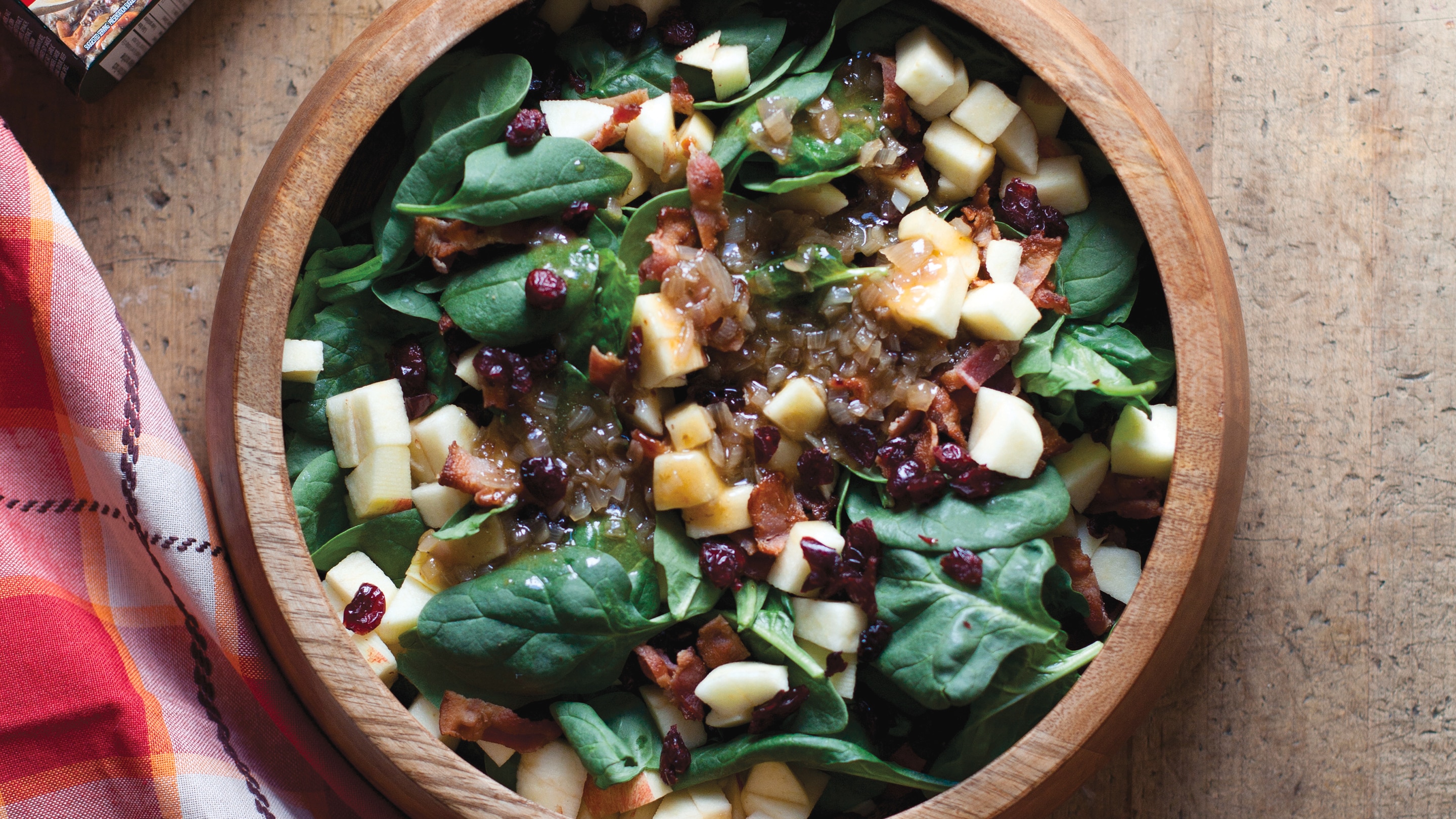 Warm spinach salad in wooden bowl covered with apple chunks, dried cranberries and dressing