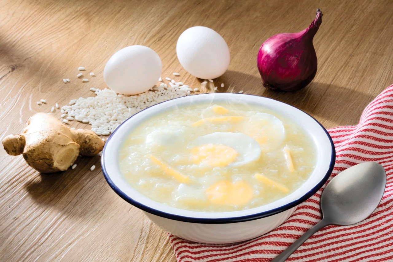 A bowl of Lugaw on a wooden surface with ginger, rice, eggs, and onions