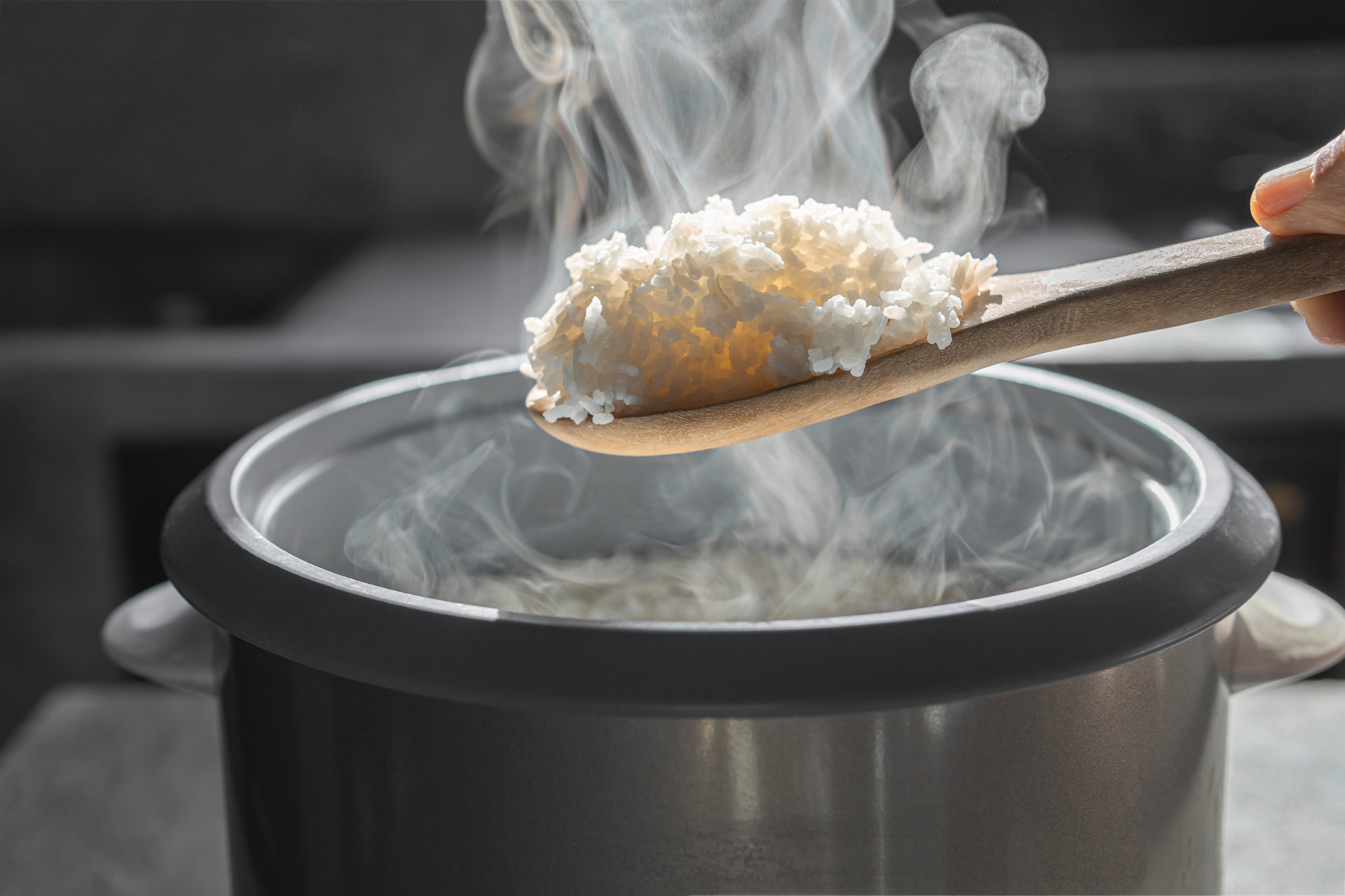 A wooden spoon filled with steaming hot rice can be seen hovering above a rice cooker appliance