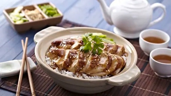 A serving of Teriyaki Chicken over rice in a clay pot, garnished with a sprinkle of sesame seeds and parsley
