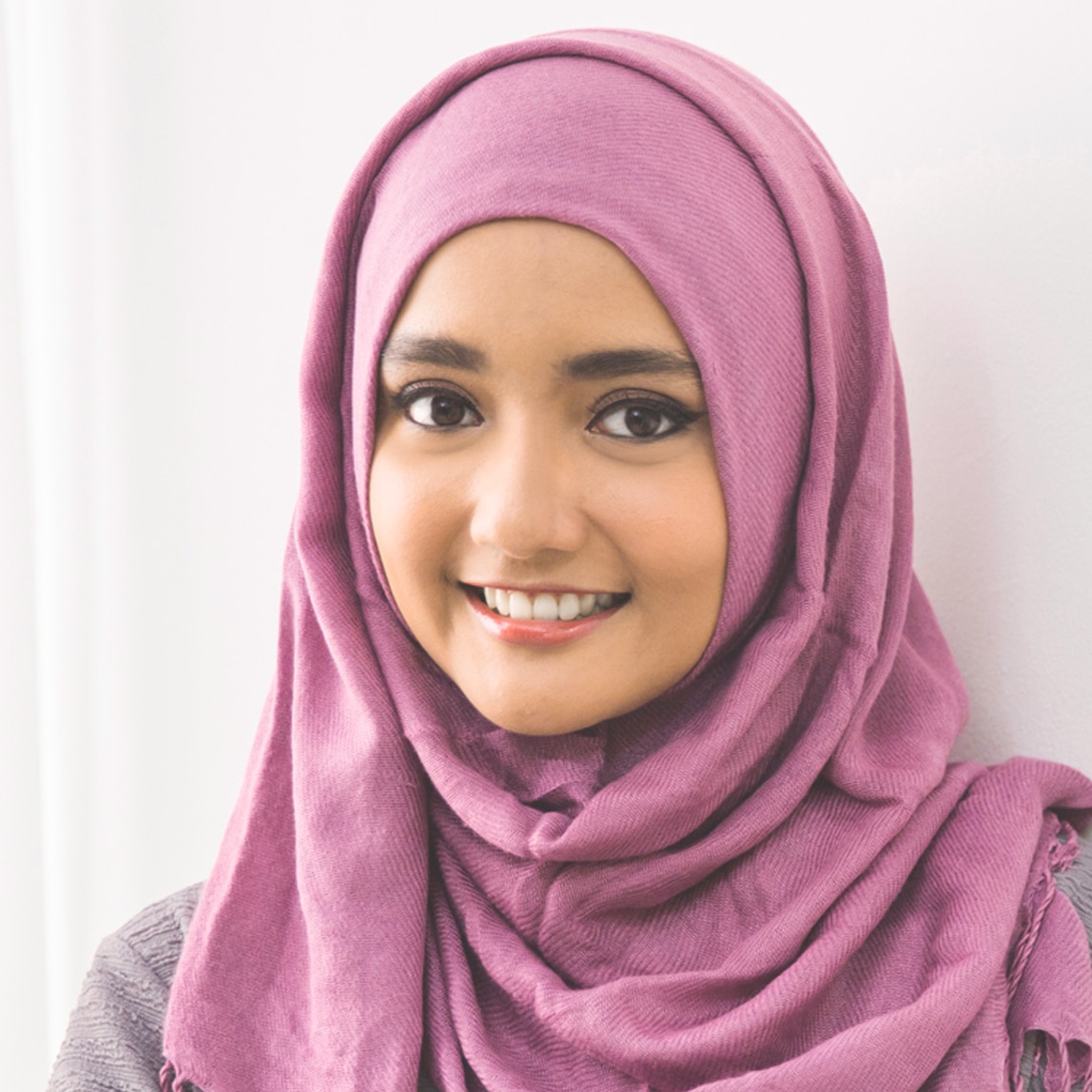 Perempuan muda mengenakan hijab ungu, tersenyum ke arah kamera.