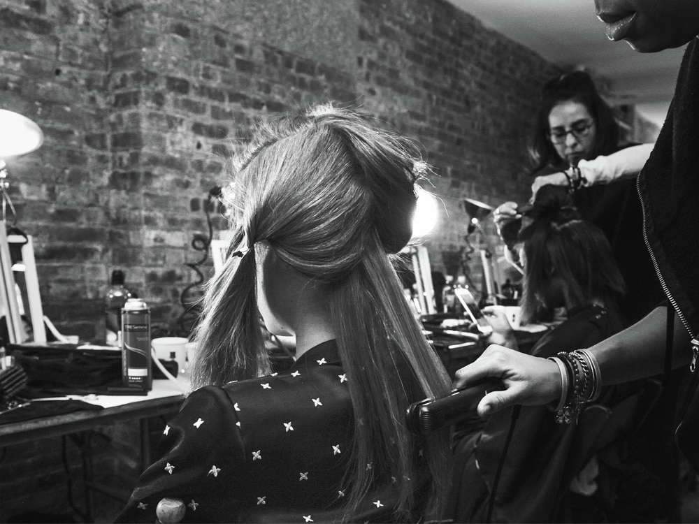 A model's head and shoulders with a stylist applying spray to her long curly hair.