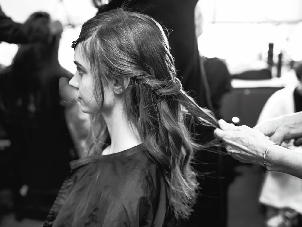 A stylist applying curling tongs to a section of a model's hair with a photographer in the background