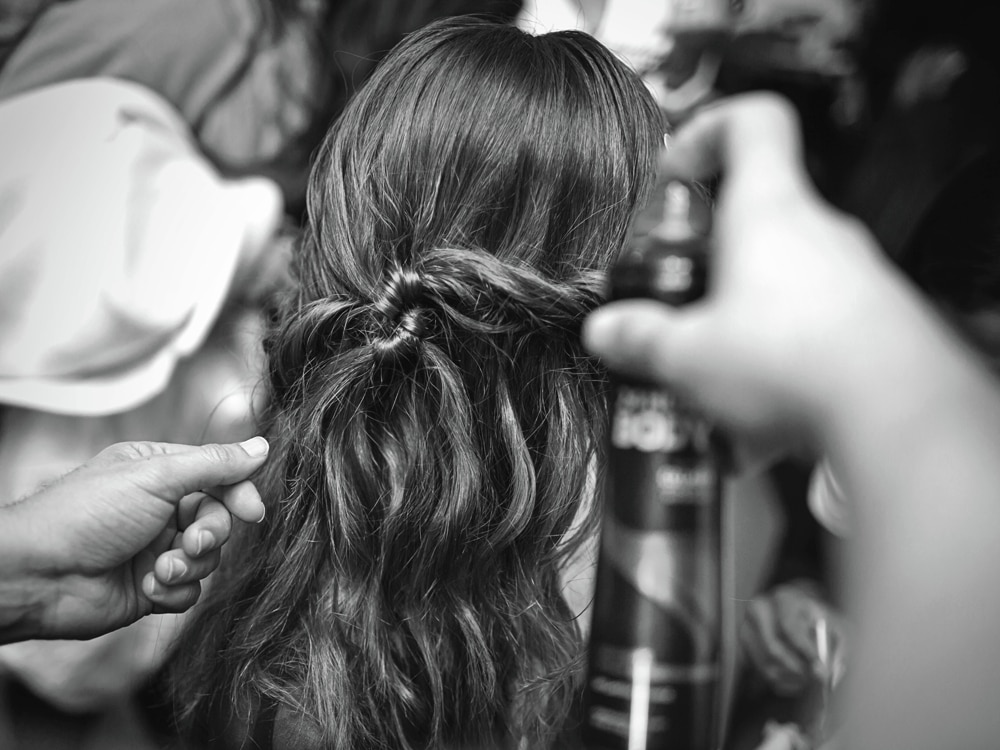 Un estilista con tenazas para rizar en una sección de cabello de una modelo y un fotógrafo en el fondo.