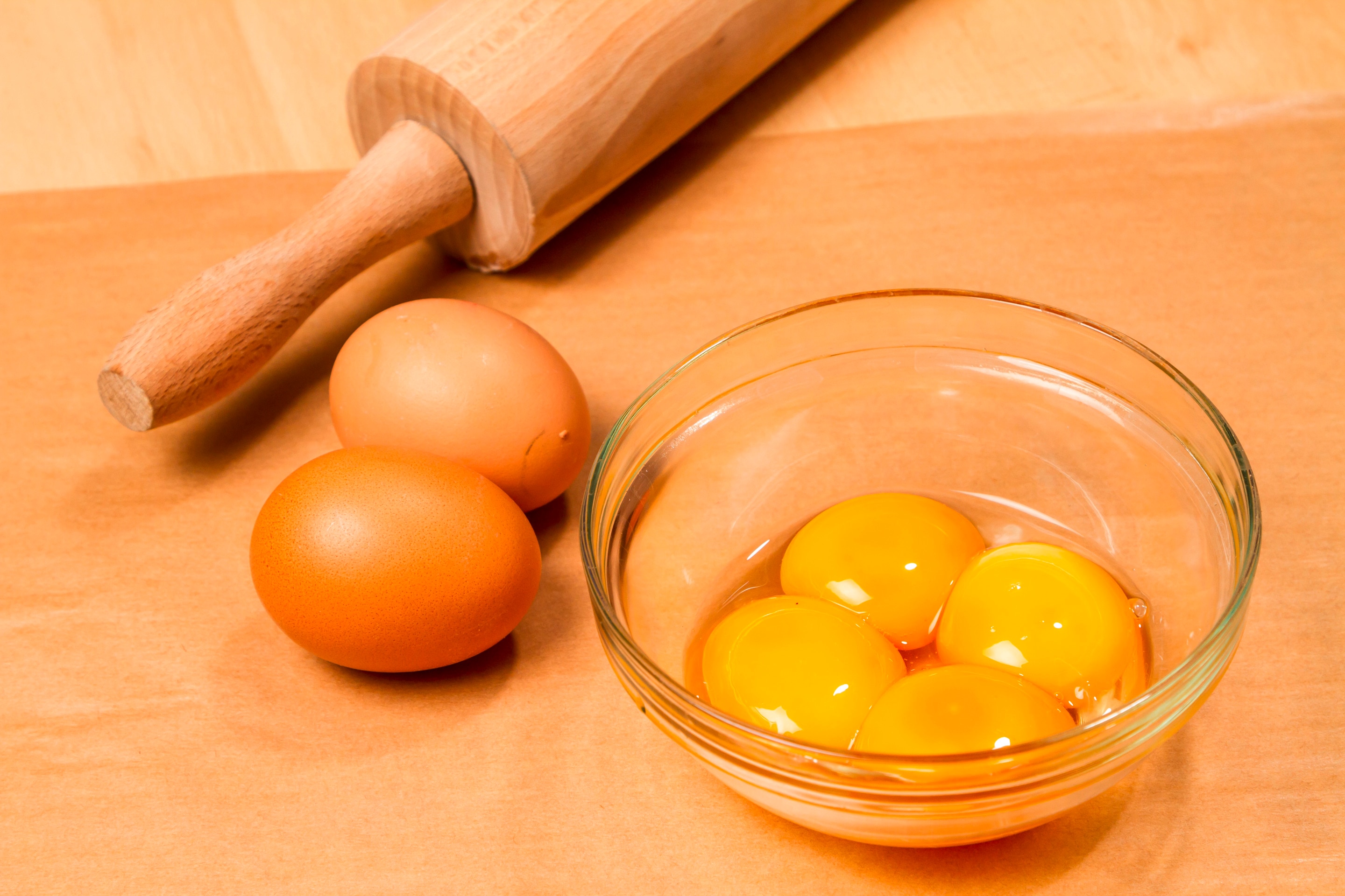 A glass bowl of 4 egg yolks next to 2 whole eggs and a rolling pin on a wooden table
