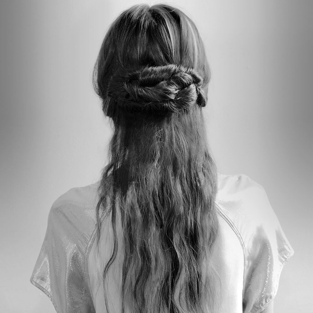 The back of a woman's head and shoulders. She wears her light brown hair in a messy, plaited bun.