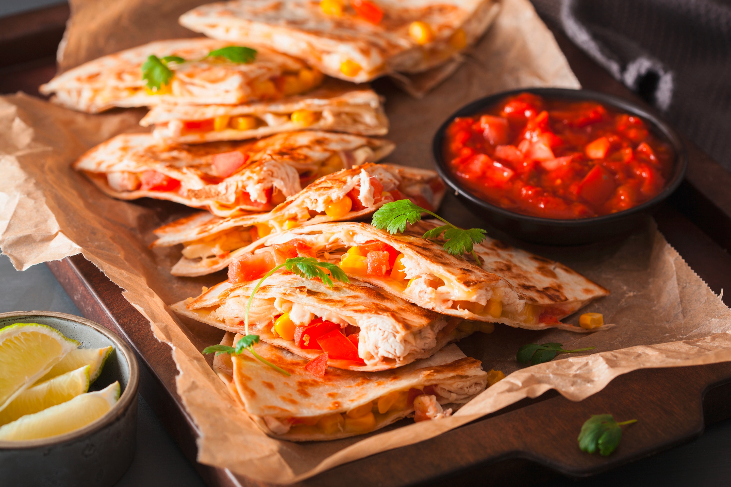 A platter of quesadillas stuffed with tomatoes, corn, cheese, and chicken, served with a side of tomato salsa and limes