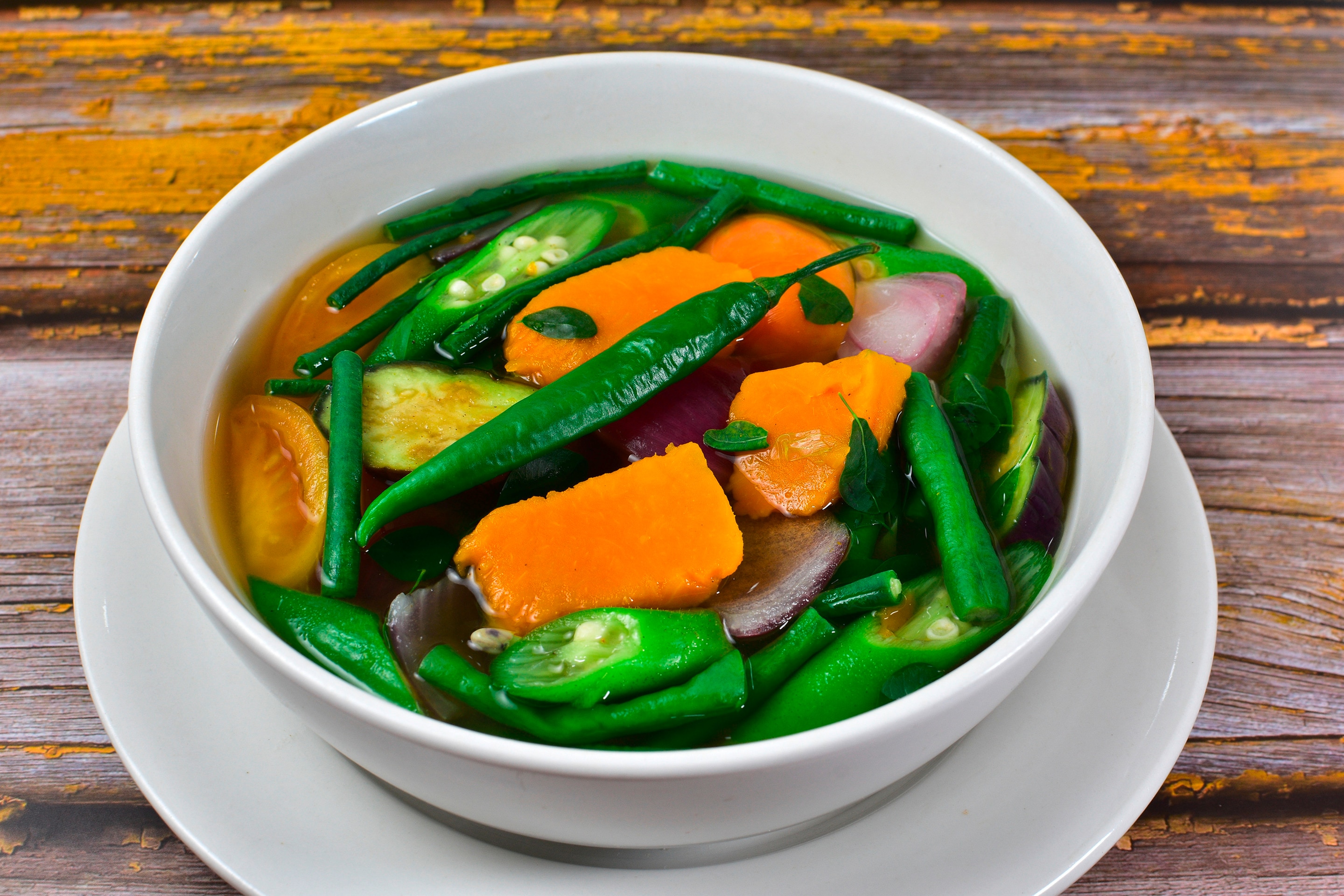 A white bowl with squash, okra, red onions, tomatoes, beans, malunggay, and green chili in clear broth