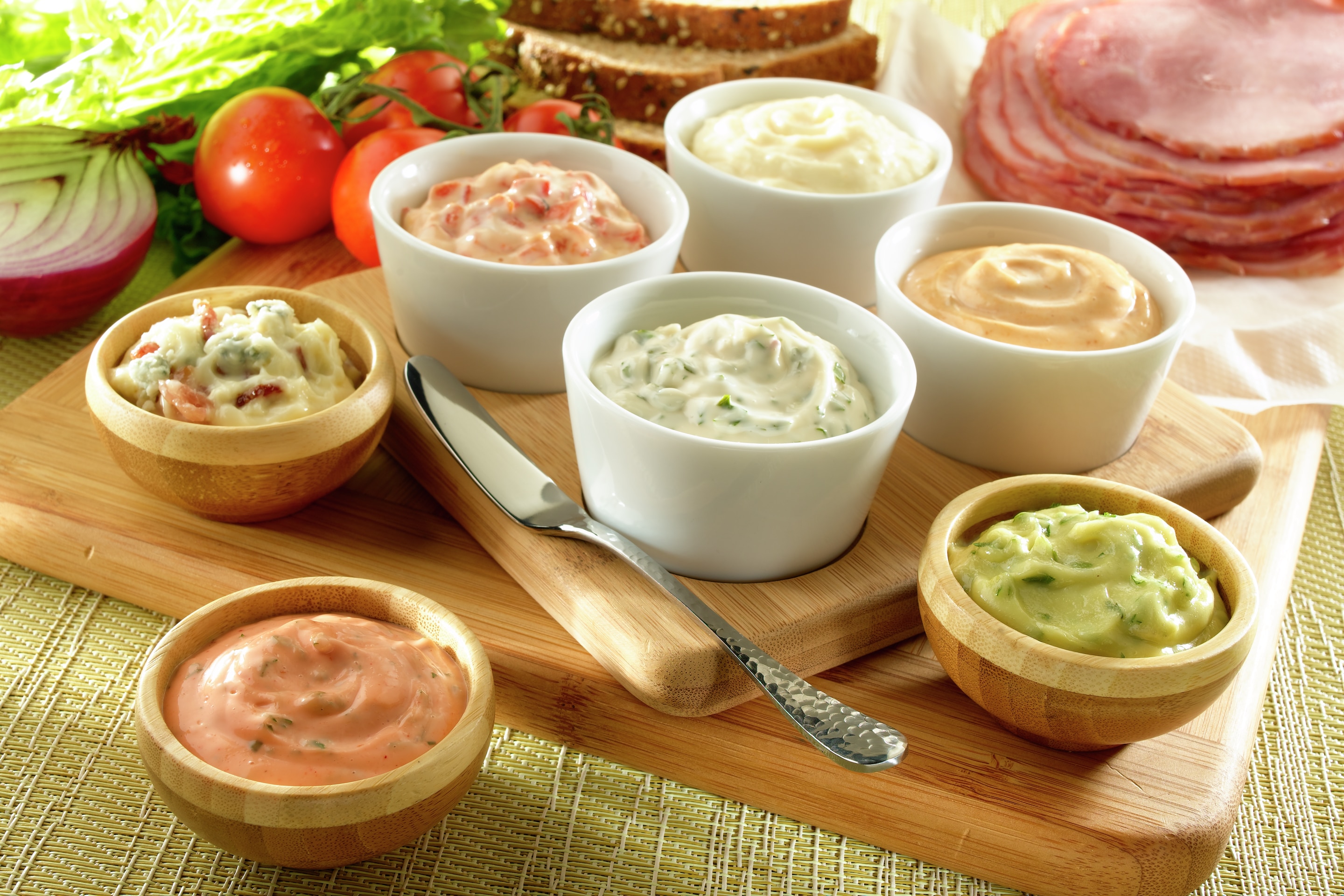 Creamy caesar spread, garlic 'N roasted red pepper spread, wasabi spread, blue cheese 'N bacon spread, buffalo spread, fresh italian herb spread, and smokey chipotle spread, served in a white bowl and placed on a wooden platform.
