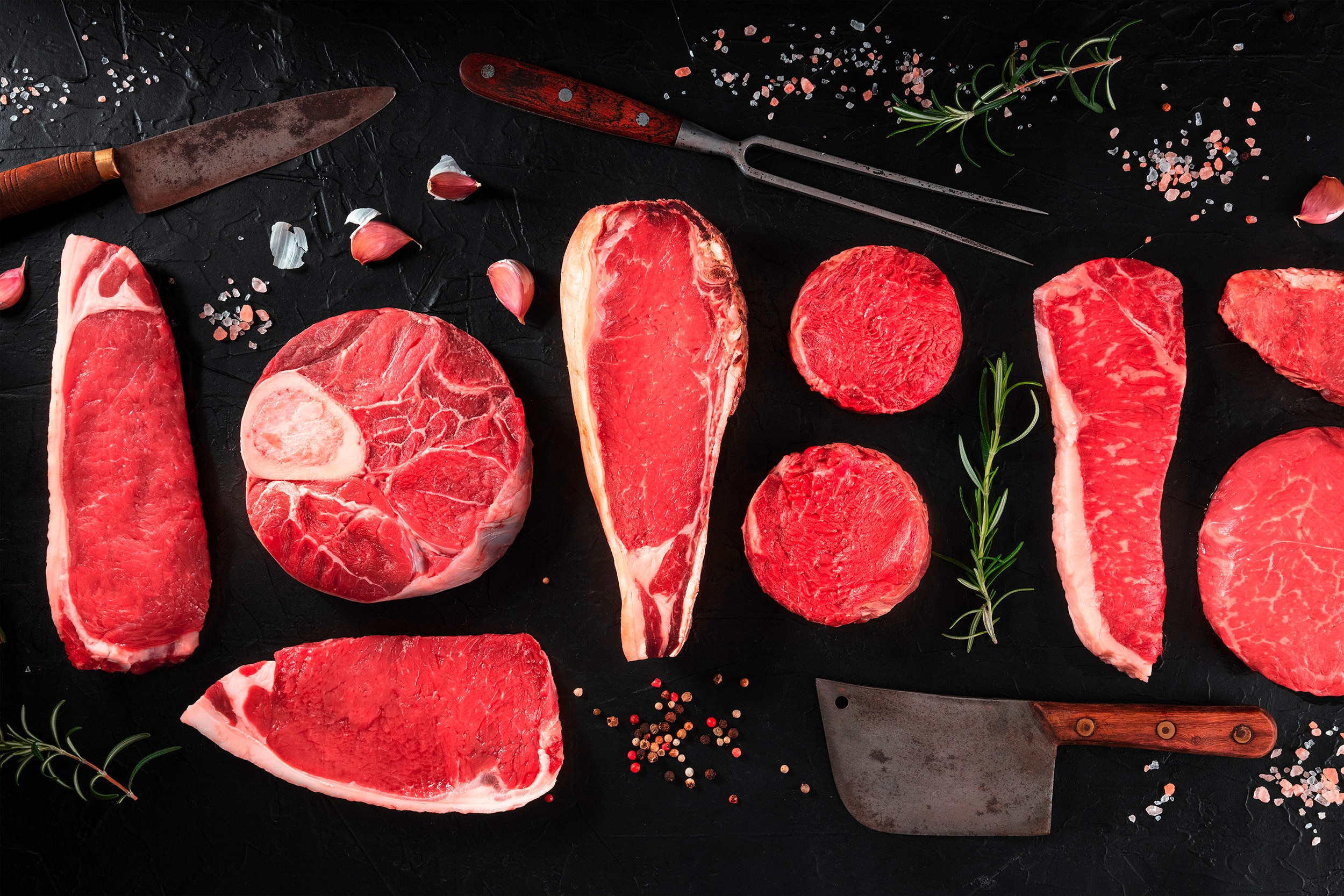 Several cuts of beef on a black countertop surrounded by different knives