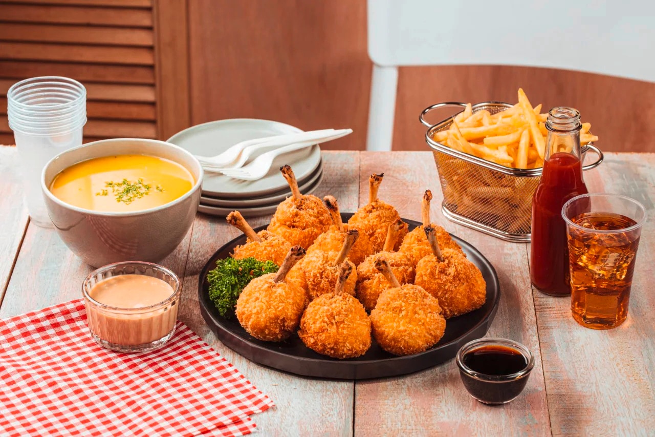 A plate of chicken lollipops, basket of fries, bowl of soup, dipping sauces, and a beverage on a wooden table