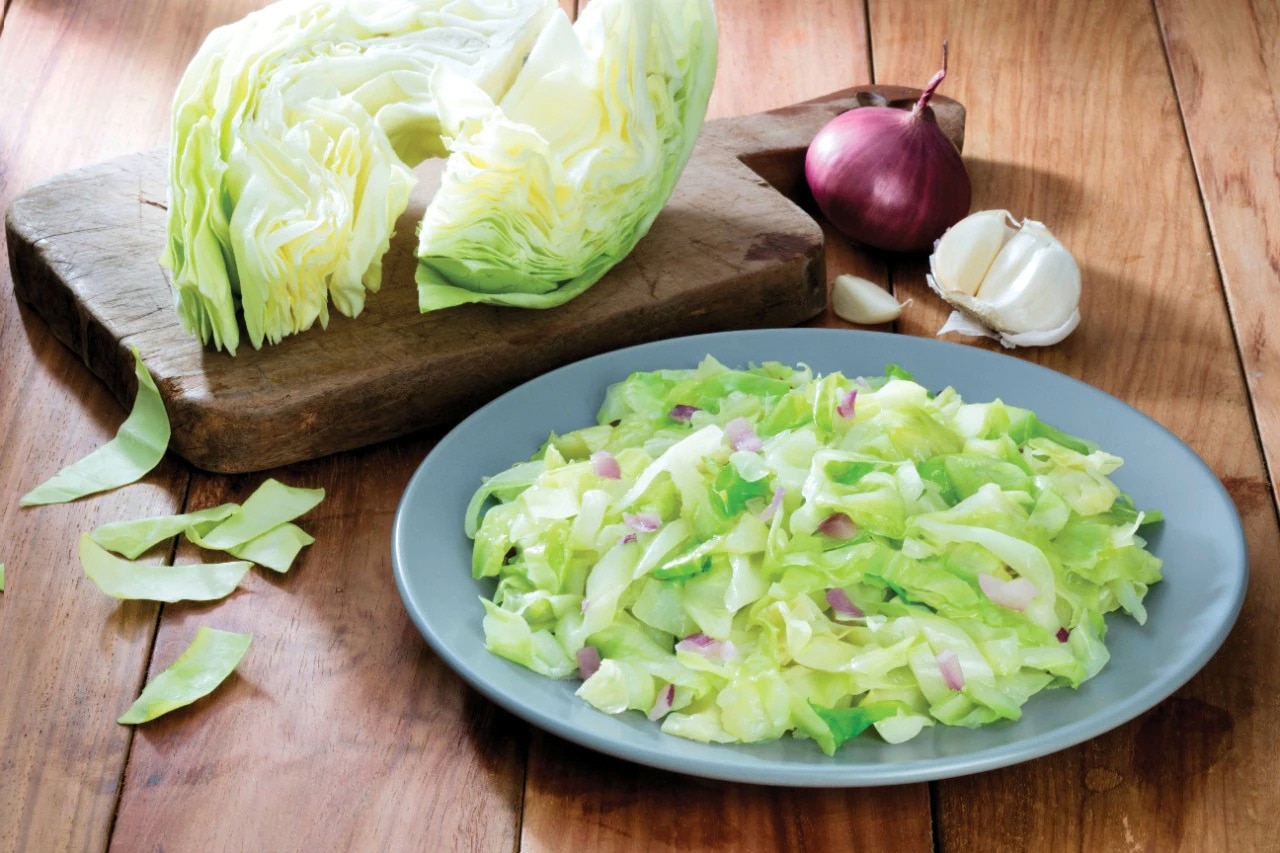 A plate of ginisang repolyo on a wooden table next to raw cabbage, garlic, and onion